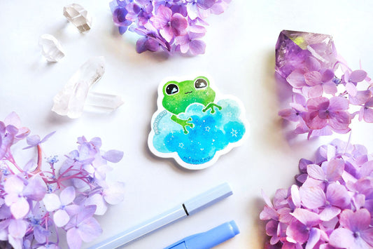 original watercolour painting by moonchu turned into a vinyl sticker! depicts a sweet green frog holding onto a sky blue cloud, with little stars twinkling and clouds in his eyes. quartz crystals and purple and blue hydrangea flowers litter the backdrop.
