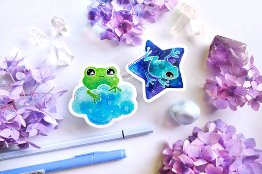 original watercolour painting by moonchu turned into a vinyl sticker! depicts a sweet green frog holding onto a sky blue cloud, with little stars twinkling and clouds in his eyes. quartz crystals and purple and blue hydrangea flowers litter the backdrop. another frog sticker friend is pictured as well. he is a teal cutie on a vibrant blue star, my personal favourite of the group.