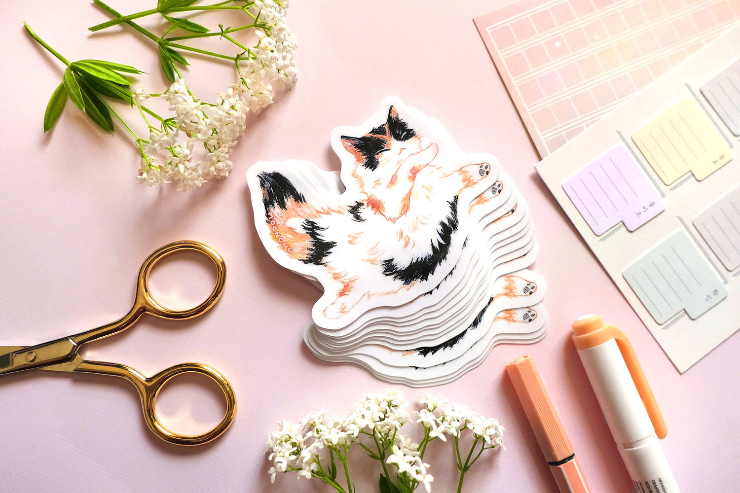 original cat illustration by moonchu depicting a sweet little calico cat baby! with orange and black patches on fluffy white fur. this cute calico kitty is floating through space with her pose. the rest of the flatlay consists of stationery items and flowers. a sweet sticker for cat lovers.