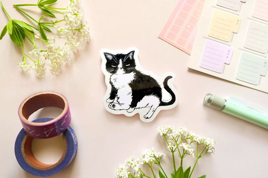 original art sticker by moonchu, illustrating a sweet little tuxedo cat. this high quality vinyl black kitty sticker is very glossy, and a tribute to mr. kitty. he is sitting with one of his signature legs stuck out. the rest of the flatlay consists of some stationery items and some flowers. a perfect sticker for tuxedo and black cat lovers!