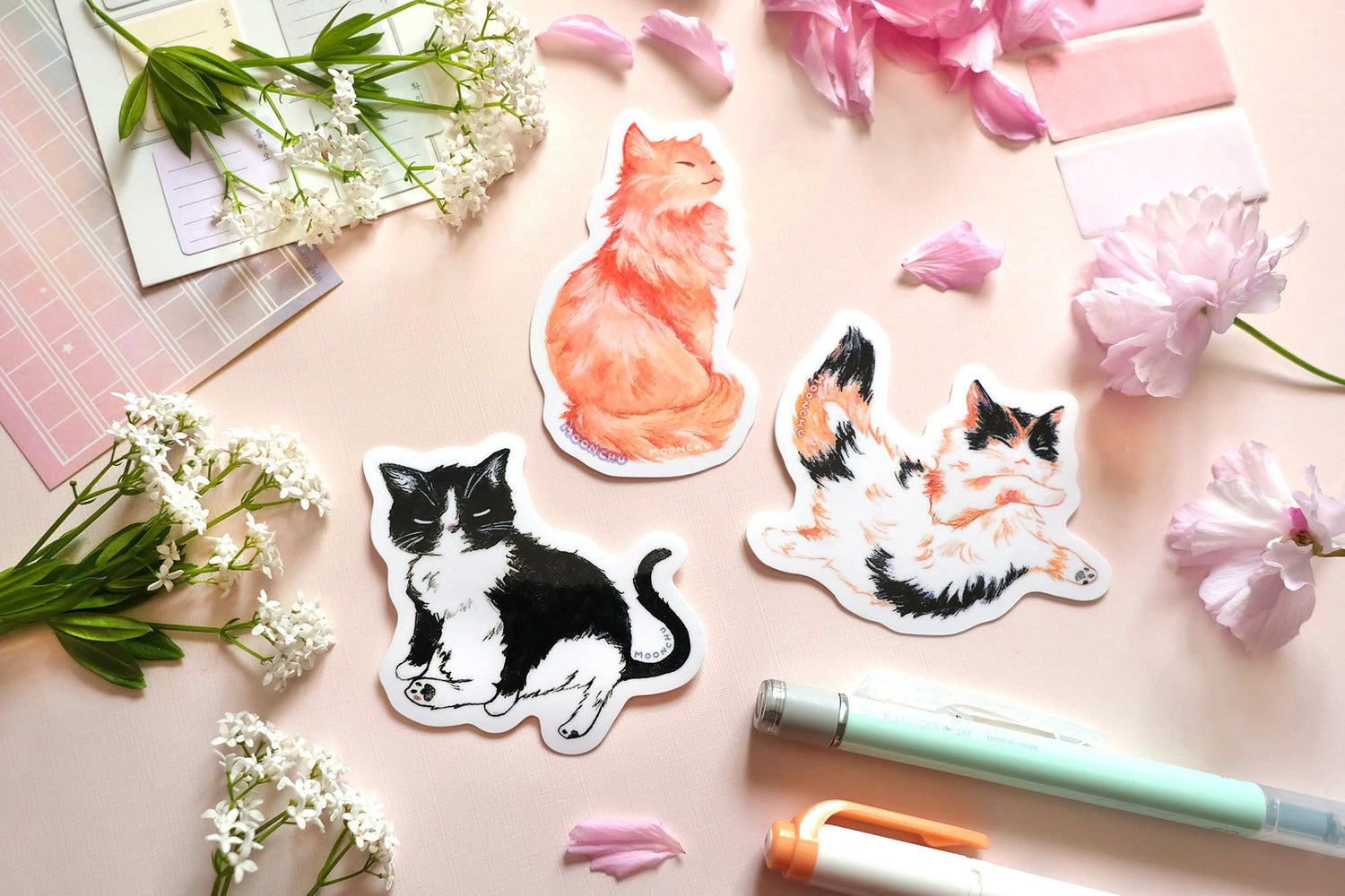 original art stickers by moonchu, illustrating a sweet little tuxedo black cat, a regal fluffy orange kitty, and a multi-coloured calico cat baby. high quality vinyl stickers, very glossy, very cute. the rest of the flatlay consists of some pink stationery items and some flowers. a perfect sticker set for all kinds of cat lovers.