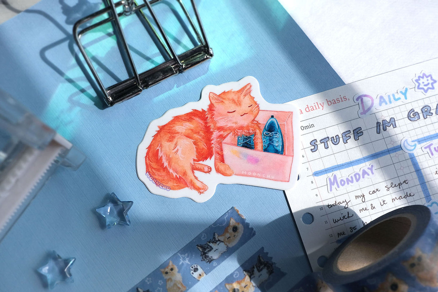 original art sticker by moonchu. vibrant high quality vinyl sticker inspired by her own cat. fluffy orange kitty painted in watercolours, lovingly holding a shoe box with blue shoes within. a very cute cat, a very glossy sticker. flatlay photo also depicts blue stationery items! including an original artist washi tape design of, you guessed it, cats by moonchu.