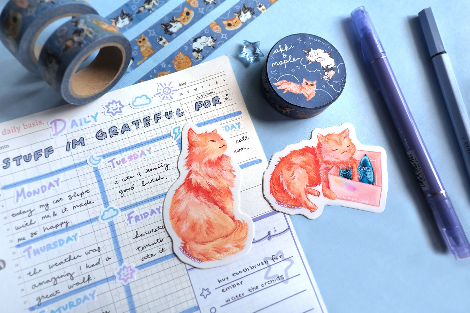 two original art stickers by moonchu. vibrant high quality vinyl stickers inspired by her own cat. fluffy orange kitty painted in watercolors, one holding a shoe box, and one sitting tall looking quite regal! a very cute cat, a very glossy sticker. flatlay photo also depicts blue stationery items! including an original artist washi tape design of, you guessed it, cats by moonchu.