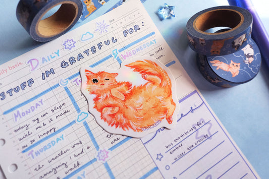original illustrated sticker by moonchu of her fluffy orange kitty! painted in watercolours, this high quality vinyl sticker illustrates a happy winky cat lying on his back. flatlay photo consists of blue stationery items, including a 'grateful' journal entry and original artist washi tape design by moonchu of, you guessed it, her cats.
