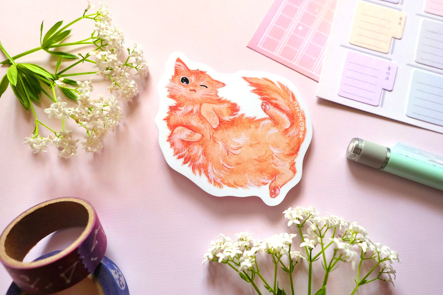 original illustrated sticker by moonchu of her fluffy orange kitty! painted in watercolours, this high quality vinyl sticker illustrates a happy winky cat lying on his back. flatlay photo consists of pink stationery items and flowers. a sweet sticker for cat lovers and stationery lovers alike.