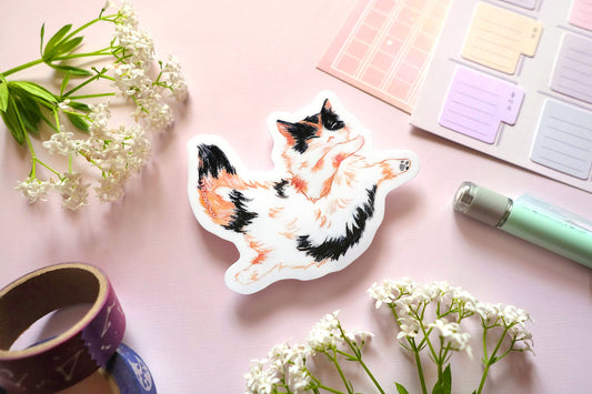 original cat illustration by moonchu depicting a sweet little calico cat baby! with orange and black patches on fluffy white fur. this cute calico kitty is floating through space with her pose. the rest of the flatlay consists of stationery items and flowers. a sweet sticker for cat lovers.