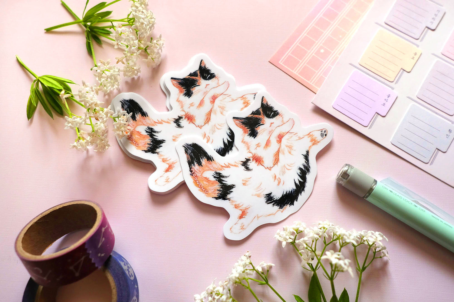 original cat illustration by moonchu depicting a sweet little calico cat baby! with orange and black patches on fluffy white fur. this cute calico kitty is floating through space with her pose. the rest of the flatlay consists of stationery items and flowers. a sweet sticker for cat lovers.