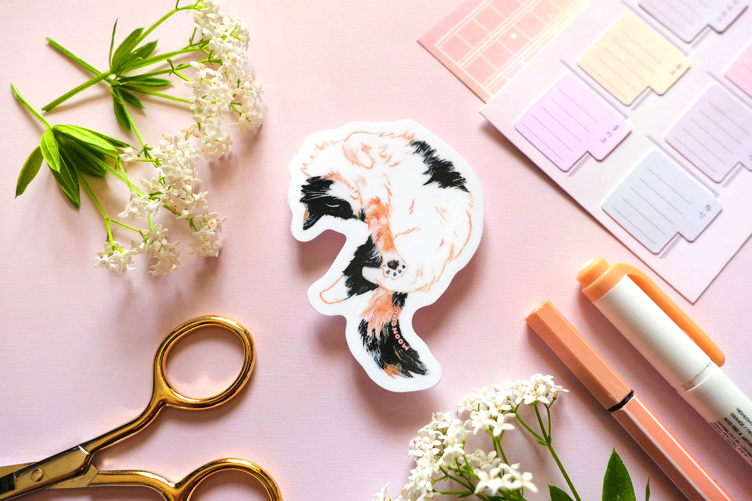 original cat illustration by moonchu depicting a sweet little calico cat baby! with orange and black patches on fluffy white fur. this cute calico kitty is having a nice stretch during a sunny cat nap. the rest of the flatlay consists of stationery items and flowers. a sweet sticker for cat lovers.
