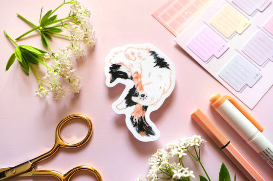 original cat illustration by moonchu depicting a sweet little calico cat baby! with orange and black patches on fluffy white fur. this cute calico kitty is having a nice stretch during a sunny cat nap. the rest of the flatlay consists of stationery items and flowers. a sweet sticker for cat lovers.