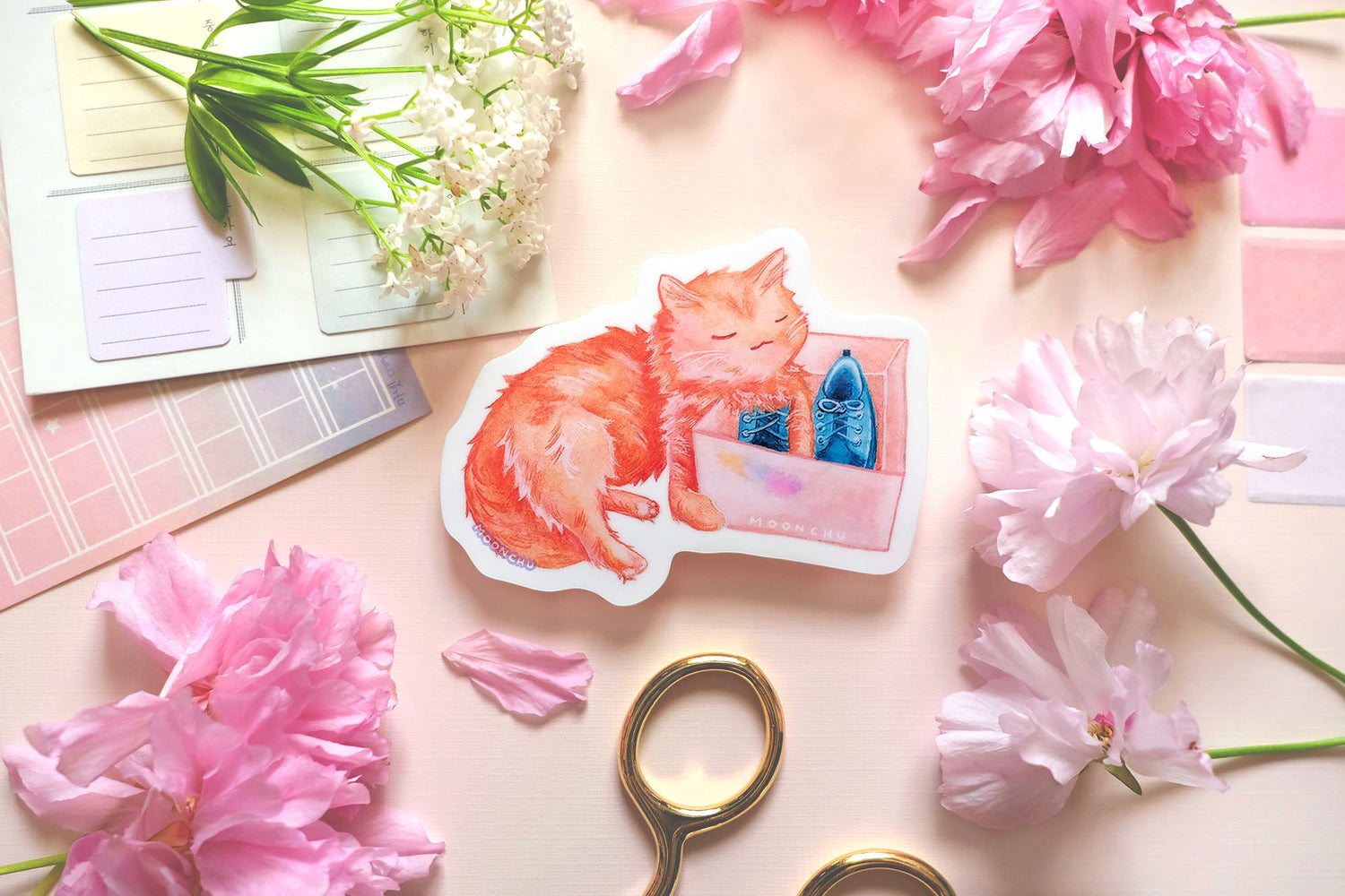 original art sticker by moonchu. vibrant high quality vinyl sticker inspired by her own cat. fluffy orange kitty watercolour sticker, lovingly holding a shoe box with blue shoes within. a very cute cat, a very glossy sticker. flatlay photo also depicts pink stationery items and flowers.