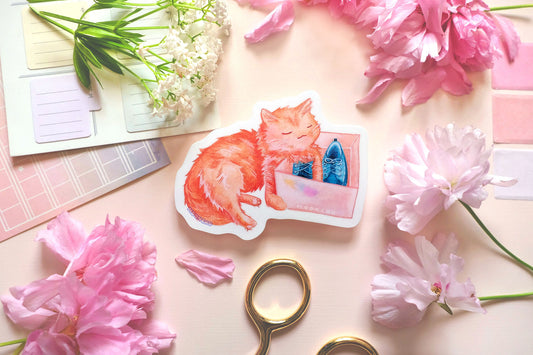 original art sticker by moonchu. vibrant high quality vinyl sticker inspired by her own cat. fluffy orange kitty watercolour sticker, lovingly holding a shoe box with blue shoes within. a very cute cat, a very glossy sticker. flatlay photo also depicts pink stationery items and flowers.