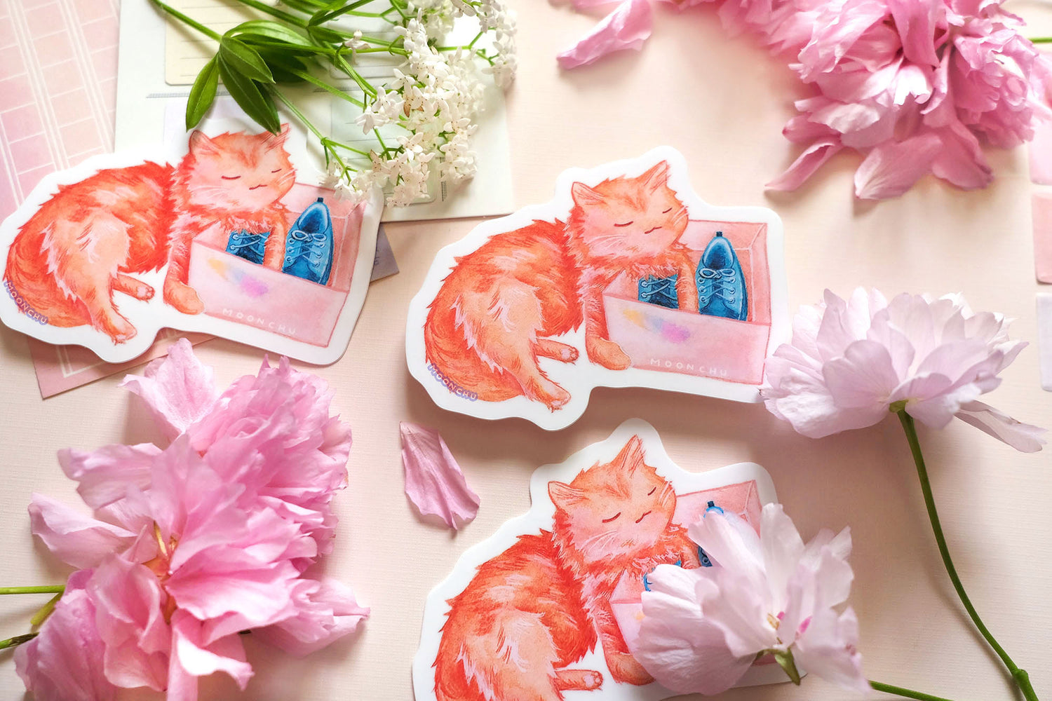 original art sticker by moonchu. vibrant high quality vinyl sticker inspired by her own cat. fluffy orange kitty watercolour sticker, lovingly holding a shoe box with blue shoes within. a very cute cat, a very glossy sticker. flatlay photo also depicts pink stationery items and flowers.
