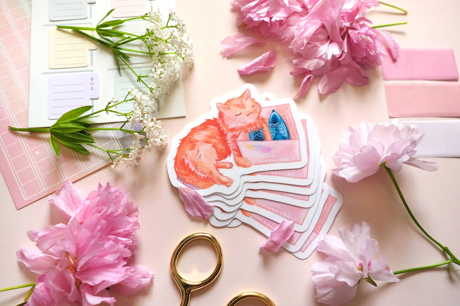 original art sticker by moonchu. vibrant high quality vinyl sticker inspired by her own cat. fluffy orange kitty watercolour sticker, lovingly holding a shoe box with blue shoes within. a very cute cat, a very glossy sticker. flatlay photo also depicts pink stationery items and flowers.