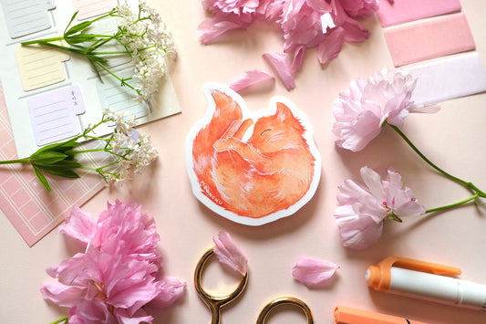 original sticker art by moonchu, watercolour orange cat painting, cat is curled up and sleepy like a donut. vibrant vinyl cat sticker is very cute, very orange! photographed on a flatlay consisting of pink stationery items and flowers.