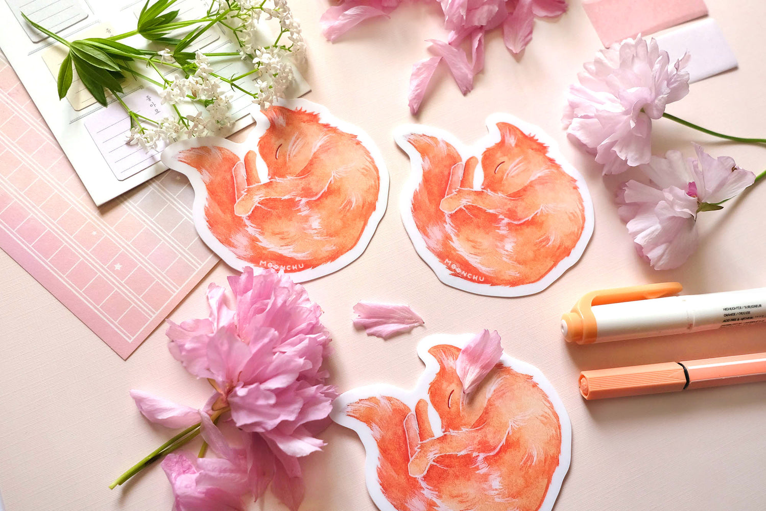 original sticker art by moonchu, watercolour orange cat painting, cat is curled up and sleepy like a donut. vibrant vinyl cat sticker is very cute, very orange! photographed on a flatlay consisting of pink stationery items and flowers.