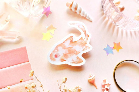 original art illustrated sticker by moonchu depicting a watercolour painted leopard shark! with a pretty pastel orange body and little constellations. has a very cute eye and curved tail. pastel flatlay consists of other ocean items, like little shells and pearls. a high quality vinyl sticker for shark lovers or ocean gifts. perfect sticker for shark week.