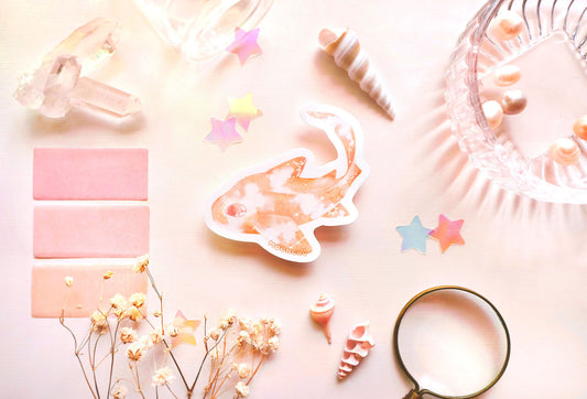 original art illustrated sticker by moonchu depicting a watercolour painted leopard shark! with a pretty pastel orange body and little constellations. has a very cute eye and curved tail. pastel flatlay consists of other ocean items, like little shells and pearls. a high quality vinyl sticker for shark lovers or ocean gifts. perfect sticker for shark week.