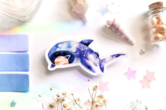 original art illustrated sticker by moonchu depicting a watercolour painted megamouth shark! with a navy blue body, it has little constellations on its body. an orange saturn sits within its notorious open mouth. flatlay consists of other ocean items, like little shells and pearls. a high quality vinyl sticker for shark lovers or ocean gifts. perfect sticker for shark week.