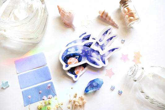 original art illustrated sticker by moonchu depicting a watercolour painted megamouth shark! with a navy blue body, it has little constellations on its body. an orange saturn sits within its notorious open mouth. flatlay consists of other ocean items, like little shells and pearls. a high quality vinyl sticker for shark lovers or ocean gifts. perfect sticker for shark week.