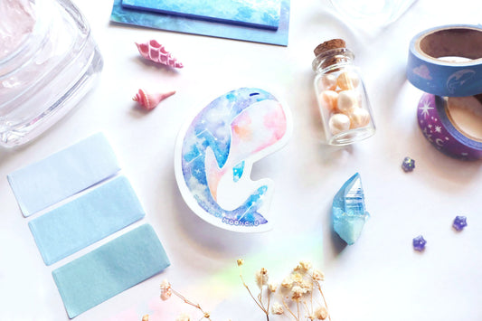 original art illustrated sticker by moonchu depicting a watercolour painted calming whale! with a serene cyan, blue, and pink gradient body and little constellations. has a sleepy face and a pink upturned belly. flatlay consists of other ocean items, like little shells and pearls. a high quality vinyl sticker for shark lovers or ocean gifts. perfect sticker for shark week.