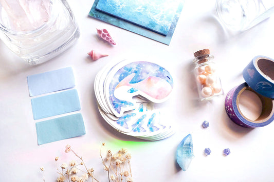original art illustrated sticker by moonchu depicting a watercolour painted calming whale! with a serene cyan, blue, and pink gradient body and little constellations. has a sleepy face and a pink upturned belly. flatlay consists of other ocean items, like little shells and pearls. a high quality vinyl sticker for shark lovers or ocean gifts. perfect sticker for shark week.