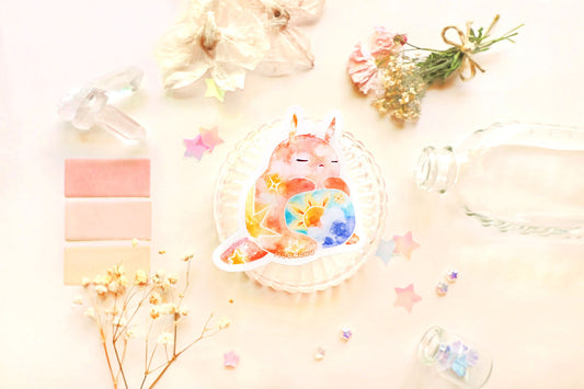 photo is of moonchu watercolor sticker depicting a v cute chubby bunny orange with yellow twinkling stars & a sun rising above blue clouds with a teal sky on his belly. he has sleepy and serene eyes. flowers, stars, and other items decorate the perimeter of sticker. a high quality colourful whimsical vinyl sticker. rest of the flatlay consists of other little pink stationery items, stars, and dried flowers.
