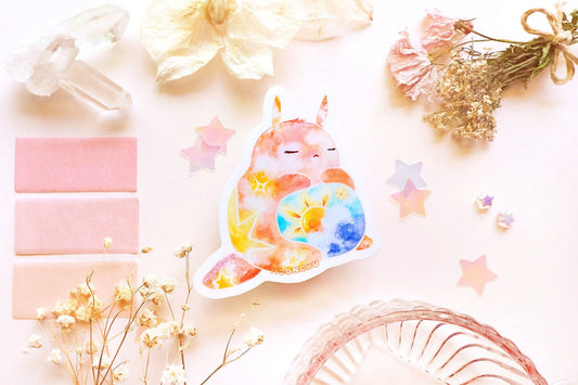 photo is of moonchu watercolor sticker depicting a v cute chubby bunny orange with yellow twinkling stars & a sun rising above blue clouds with a teal sky on his belly. he has sleepy and serene eyes. flowers, stars, and other items decorate the perimeter of sticker. a high quality colourful whimsical vinyl sticker. rest of the flatlay consists of other little pink stationery items, stars, and dried flowers.