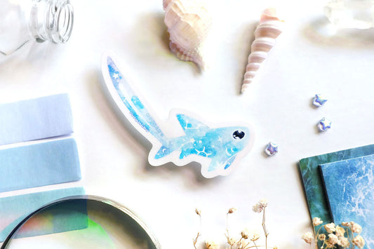 original art illustrated sticker by moonchu depicting a watercolour painted thresher shark! with a light blue body, it has little constellations on its body and a shooting star on its notoriously large tail. flatlay consists of other ocean items, like little shells and pearls. a high quality vinyl sticker for shark lovers or ocean gifts. perfect sticker for shark week.