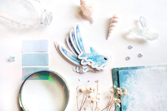 original art illustrated sticker by moonchu depicting a watercolour painted thresher shark! with a light blue body, it has little constellations on its body and a shooting star on its notoriously large tail. flatlay consists of other ocean items, like little shells and pearls. a high quality vinyl sticker for shark lovers or ocean gifts. perfect sticker for shark week.
