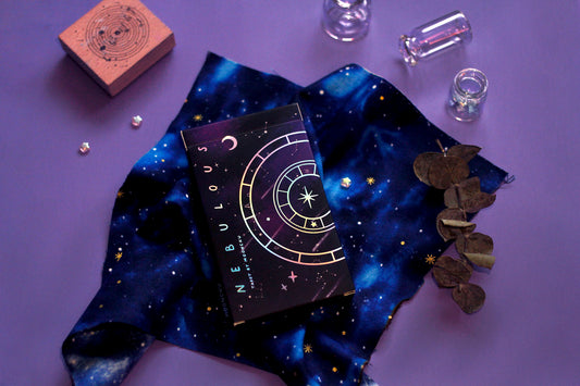 stunning indie holographic tarot deck by moonchu with all original art designs! this photograph depicts the holographic detailed tarot box design, with a galactic theme of a rich purple background and shooting stars originally painted in watercolour. this is accented by graphic designs of a crescent moon, nebulous tarot major arcana name, and intricate wheel design - all in holographic foil. comes lovingly hand-wrapped in a blue galaxy space themed cloth and twine string.